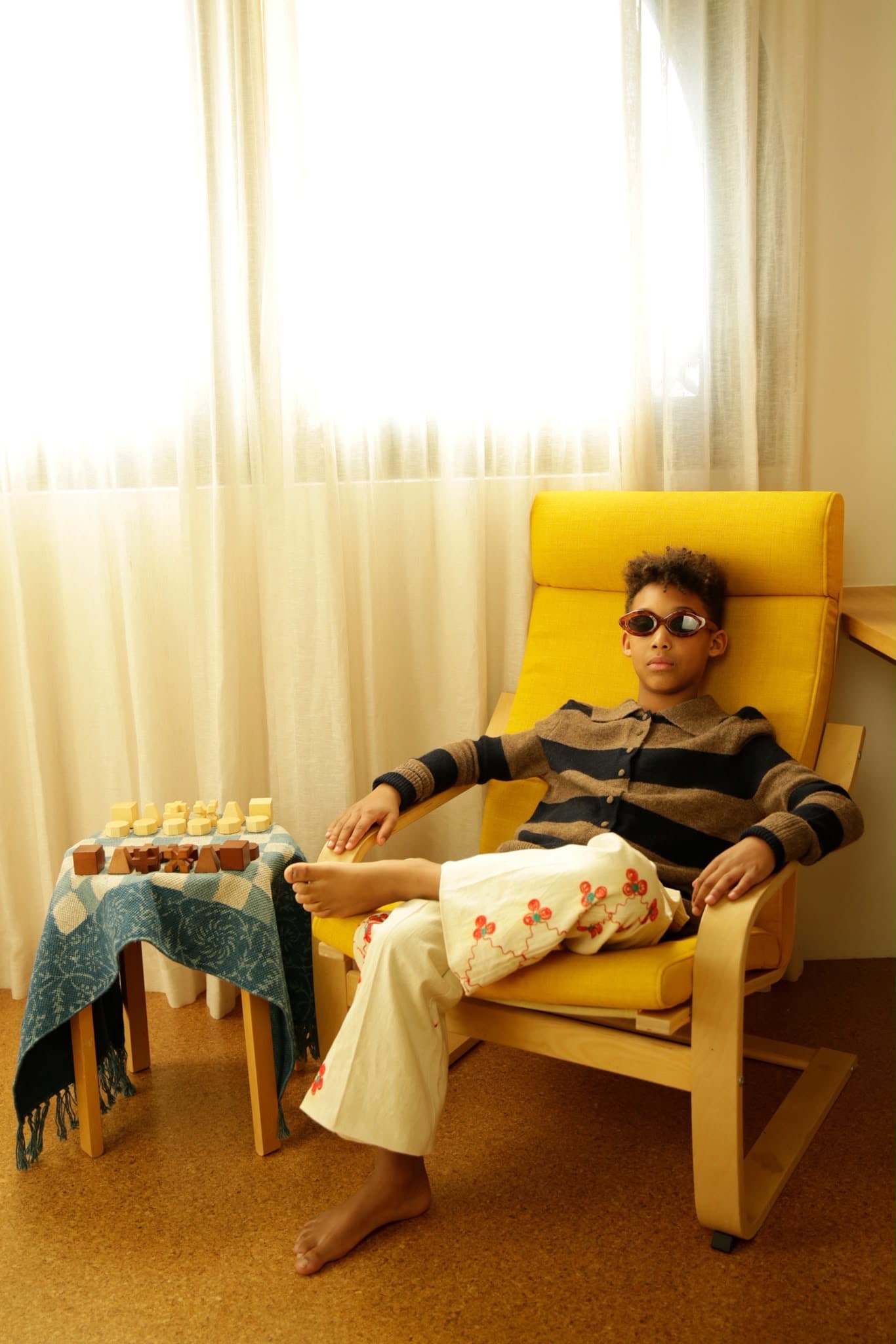 Arlo seated in a yellow chair beside a wooden chess set, backlit by a curtained window